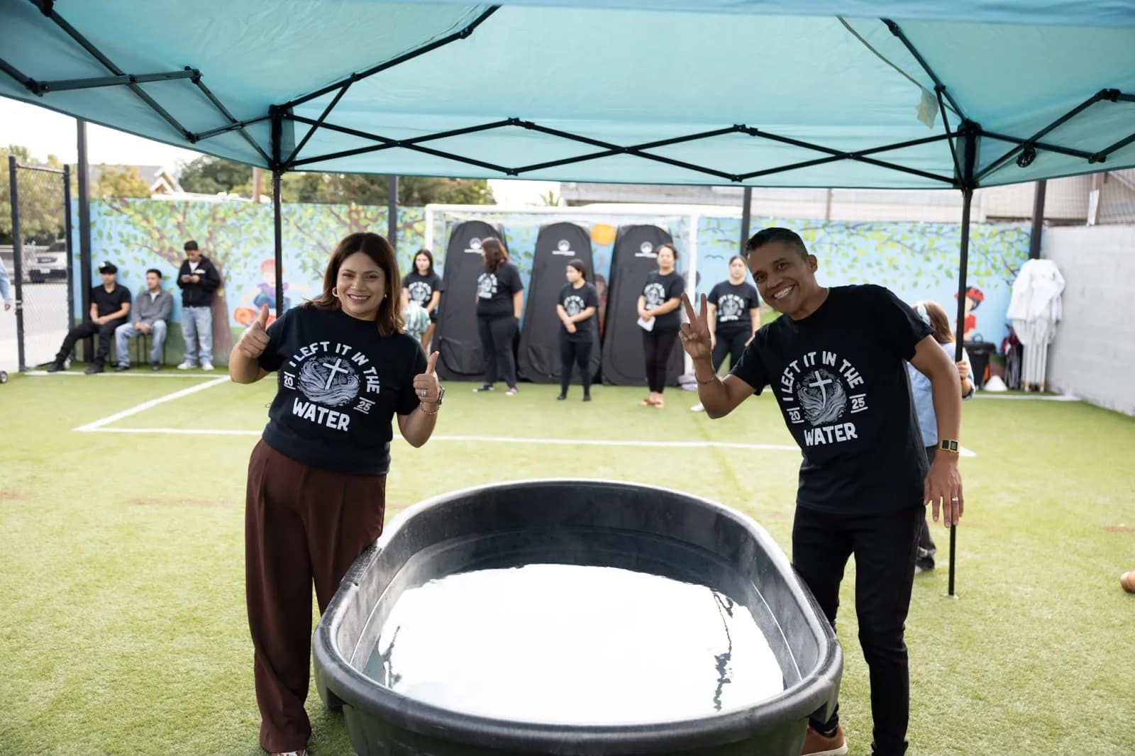 Los dos que van a bautizar levantan el pulgar junto a la pila llena de agua bajo la carpa