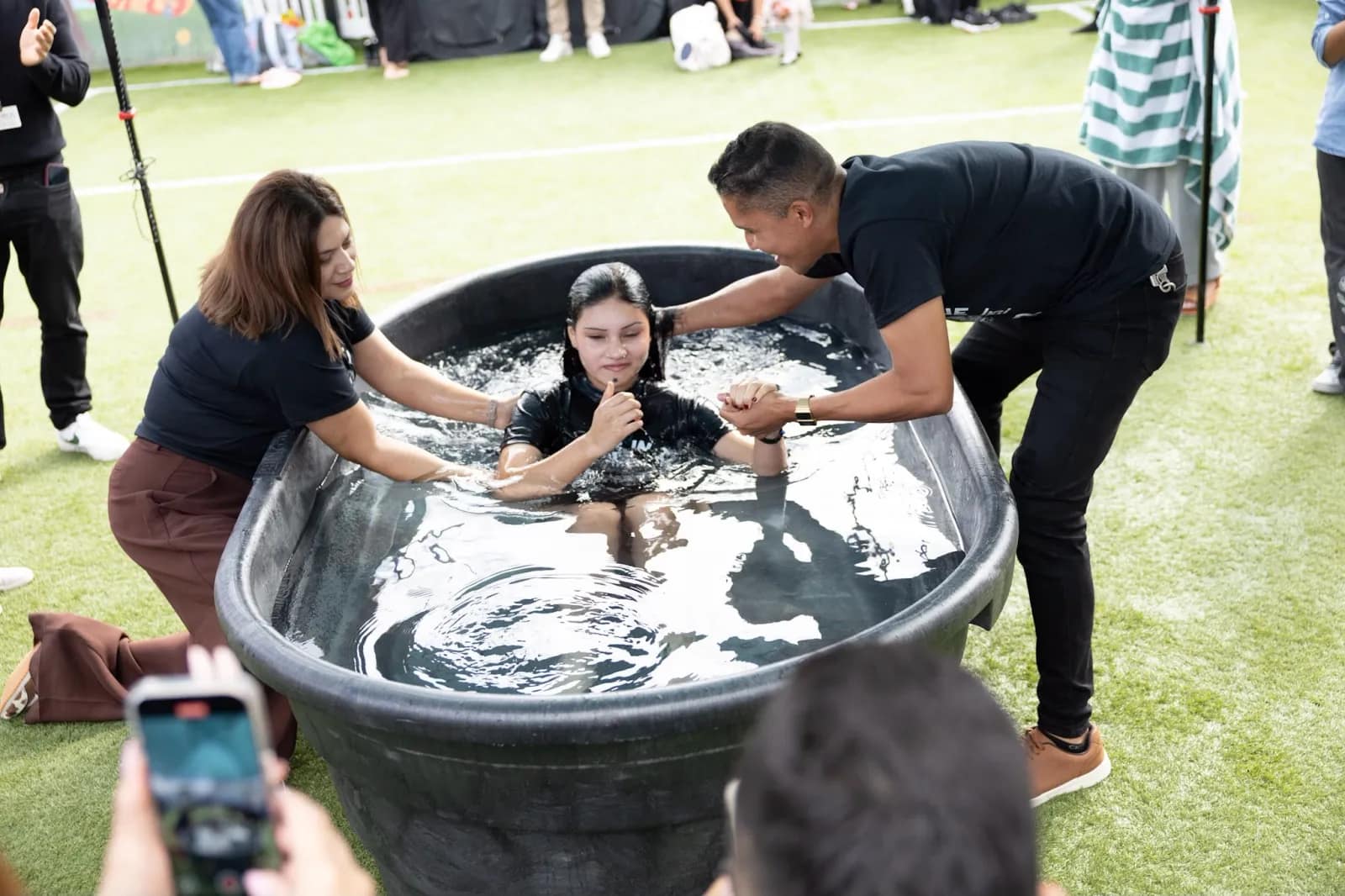 Una joven sale del agua con las manos juntas, sostenida por los brazos de los que la bautizan