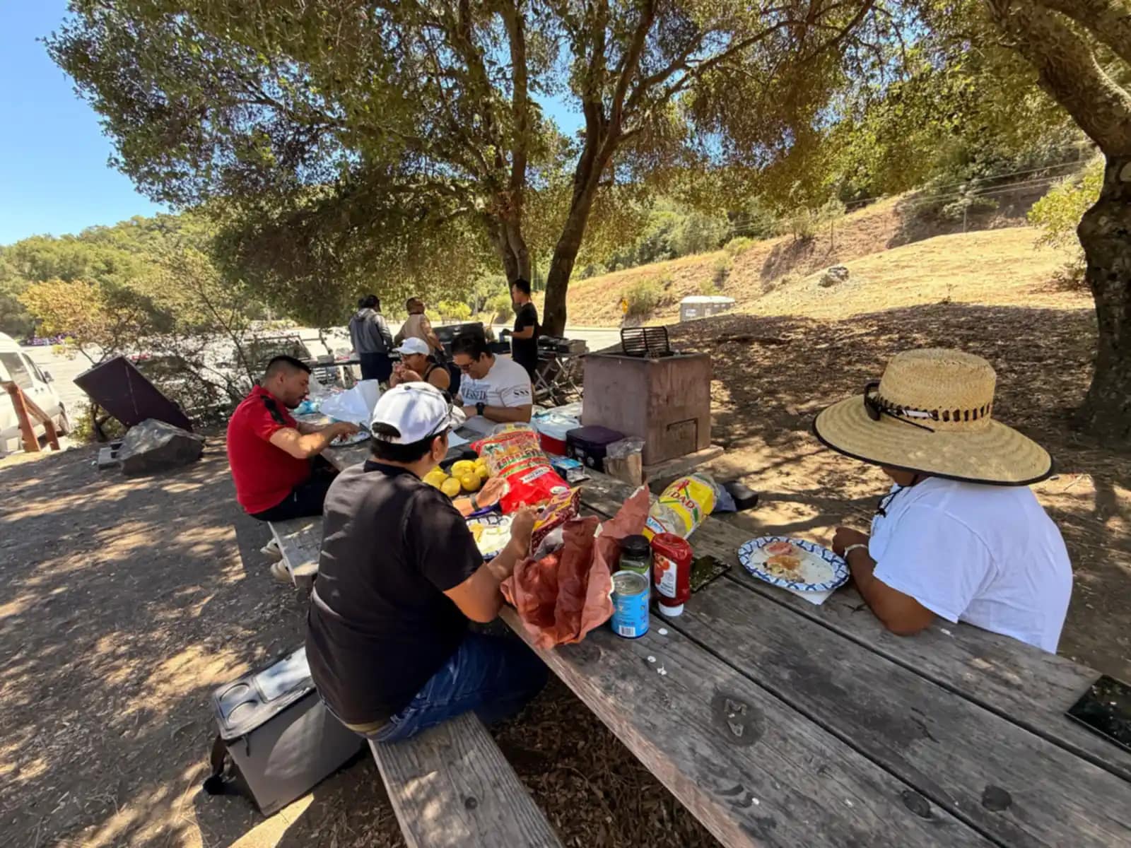 Varios hombres almuerzan en la mesa de picnic a la sombra, uno con sombrero de paja