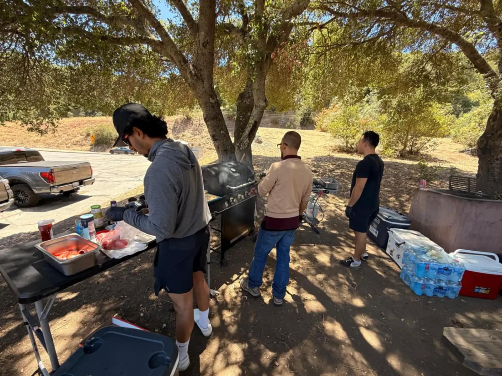 Cuatro hombres alrededor de la parrilla y la mesa de preparación, a la sombra del roble