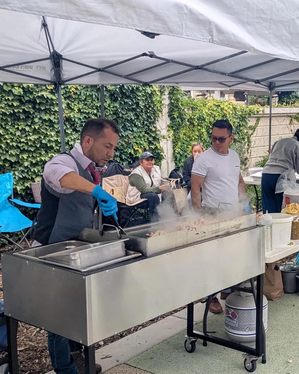 Un voluntario de Kairos cocina en una parrilla bajo una carpa en el parque, con vecinos alrededor esperando su plato